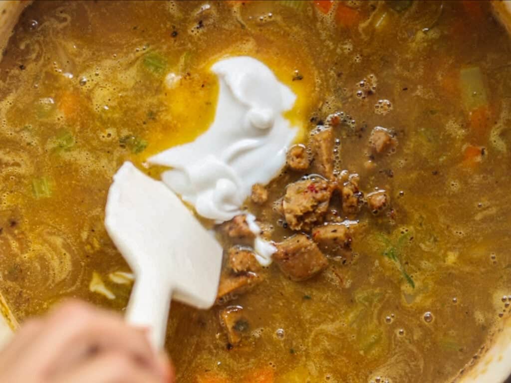 A close-up of a hand using a white spatula to stir creamy vegan yogurt and chunks of vegan sausage into a pot of brown soup with visible herbs and vegetables.